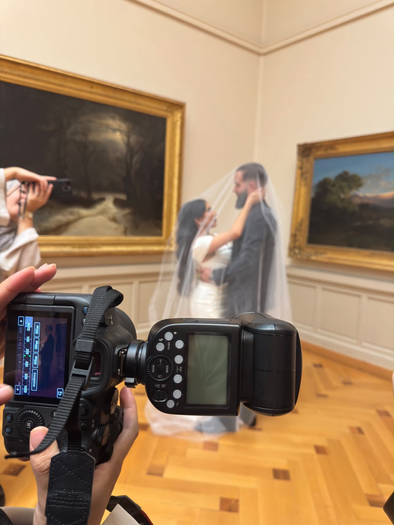 Les mariés enveloppés sous un voile de tulle, photographiés par un photographe au premier plan dans une galerie aux cadres dorés